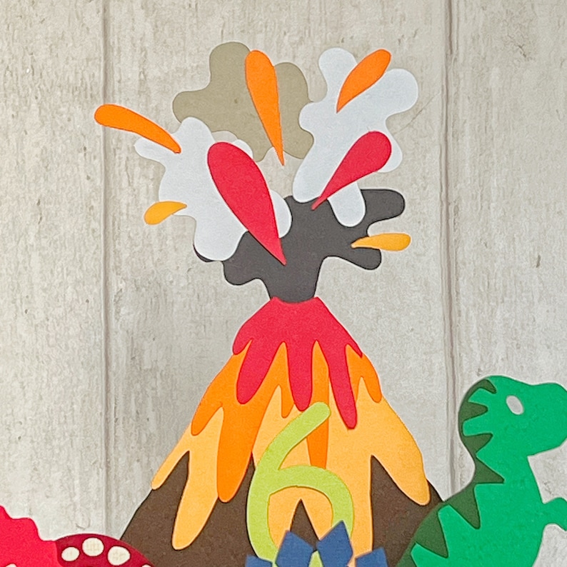 May include: A brown volcano with orange and red lava erupting. The volcano is surrounded by a white, orange, and grey cloud. A green dinosaur is on the right side of the image. The number 6 is in front of the volcano.