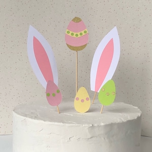 Puede incluir: Un pastel blanco decorado con adornos de pastel de huevos de Pascua rosas y verdes y dos orejas de conejo rosas. Los huevos tienen detalles de purpurina dorada.