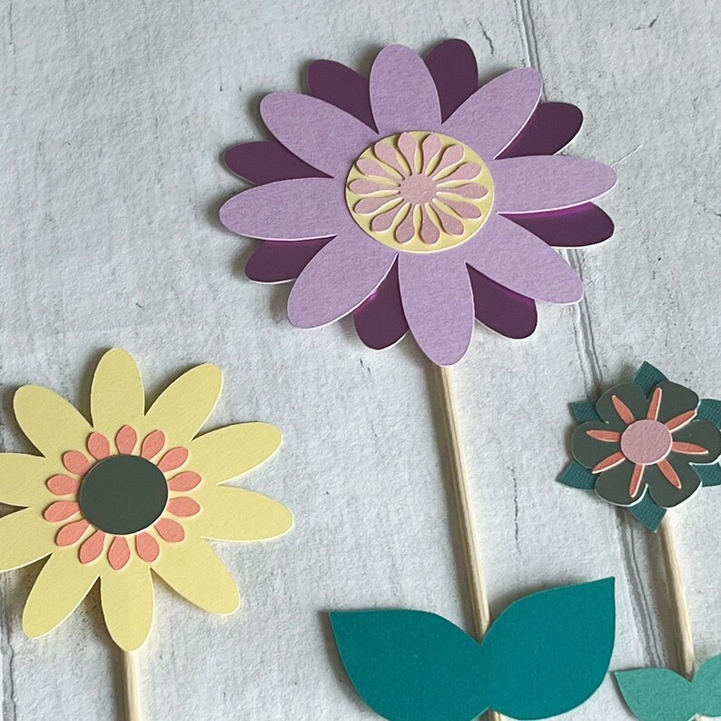 Pu&ograve; includere: Tre topper di fiori di carta con bastoncini di legno. I fiori sono viola, gialli e verdi. Il fiore viola ha un centro rosa. Il fiore giallo ha un centro arancione. Il fiore verde ha un centro rosa.