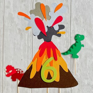 May include: A paper craft volcano with red and orange flames erupting from the top. The volcano is decorated with a green number six. A green dinosaur and a red dinosaur are on either side of the volcano.