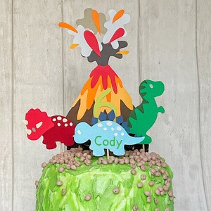 May include: A green, blue and red dinosaur cake topper with a volcano and the name Cody. The cake topper is on a green frosted cake with brown sprinkles.