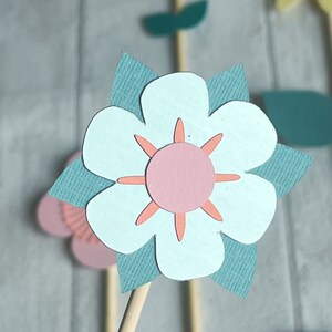 Pu&ograve; includere: Un fiore di carta blu chiaro con un centro rosa e petali di carta verde acqua. Il fiore &egrave; su un bastoncino di legno.