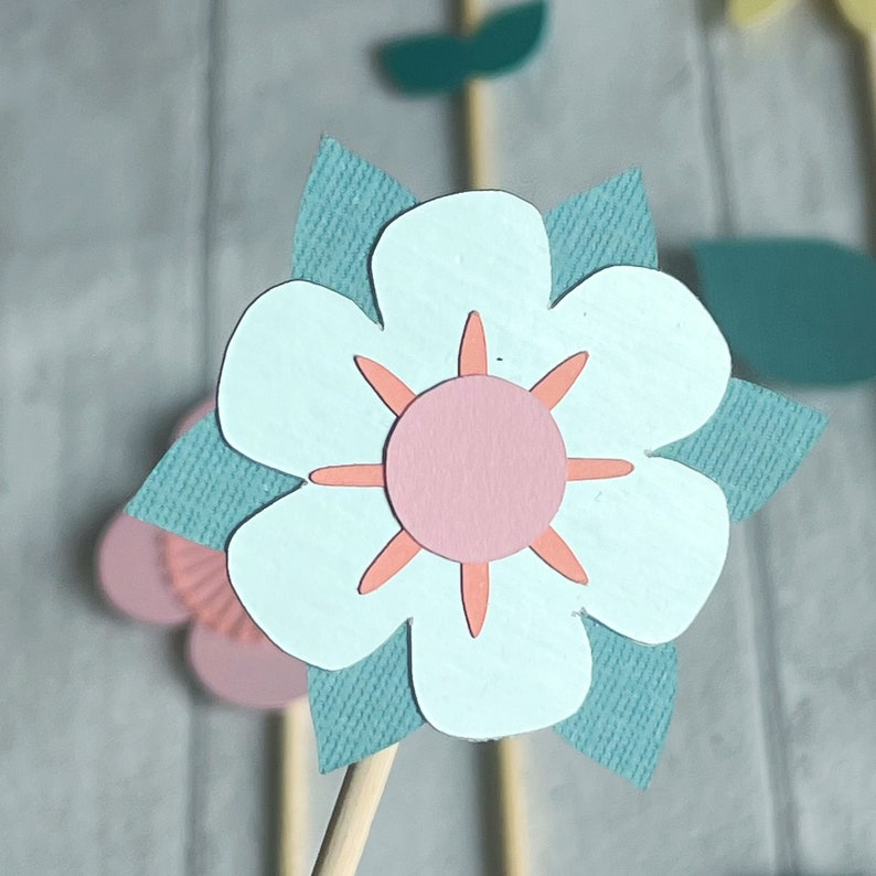 Pu&ograve; includere: Un fiore di carta blu chiaro con un centro rosa e petali di carta verde acqua. Il fiore &egrave; su un bastoncino di legno.