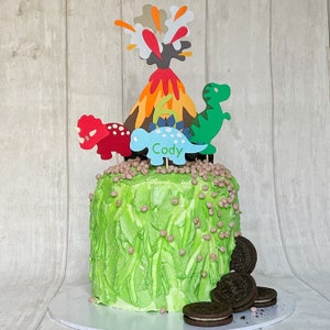 May include: Green frosted cake decorated with dinosaur and volcano themed paper cutouts. The cake is topped with chocolate chip cookies and the name "Cody" is written on one of the dinosaurs.