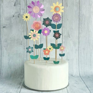 Pu&ograve; includere: Una torta bianca decorata con fiori di carta di vari colori, tra cui rosa, viola, giallo e verde. I fiori sono disposti in un design a bouquet sulla parte superiore della torta.