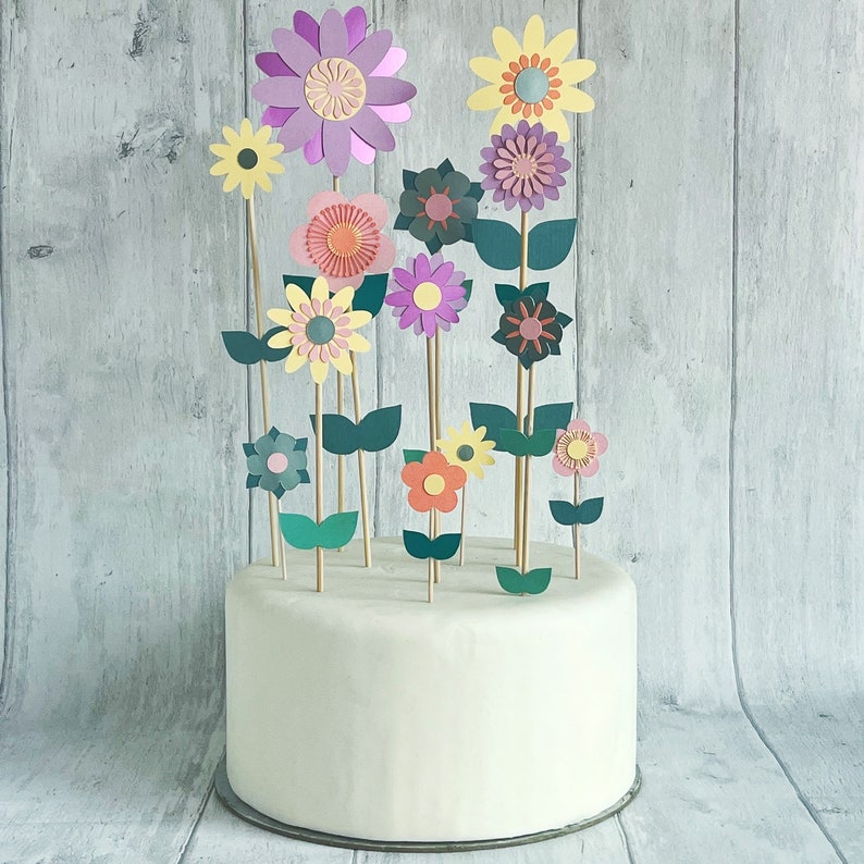 Pu&ograve; includere: Una torta bianca decorata con fiori di carta di vari colori, tra cui rosa, viola, giallo e verde. I fiori sono disposti in un design a bouquet sulla parte superiore della torta.