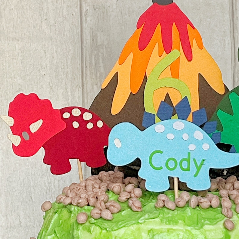 May include: A green frosted cake decorated with a volcano, two dinosaurs, and brown sprinkles. The blue dinosaur has the name "Cody" written on it.