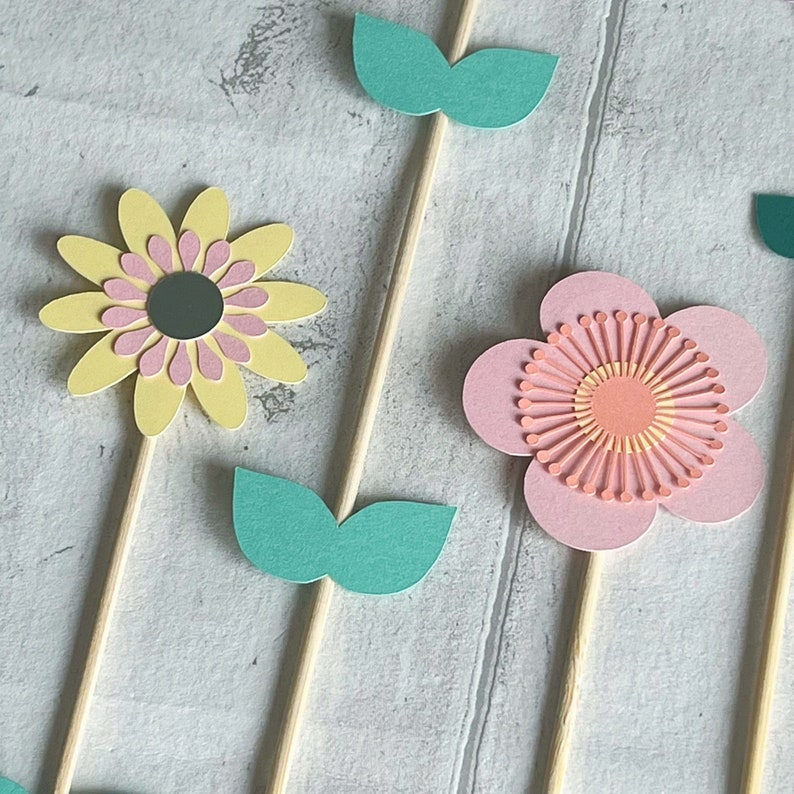 Pu&ograve; includere: Tre topper di fiori di carta con bastoncini di legno. I fiori sono gialli, rosa e pesca con foglie verdi. I fiori sono sovrapposti e hanno un effetto 3D.