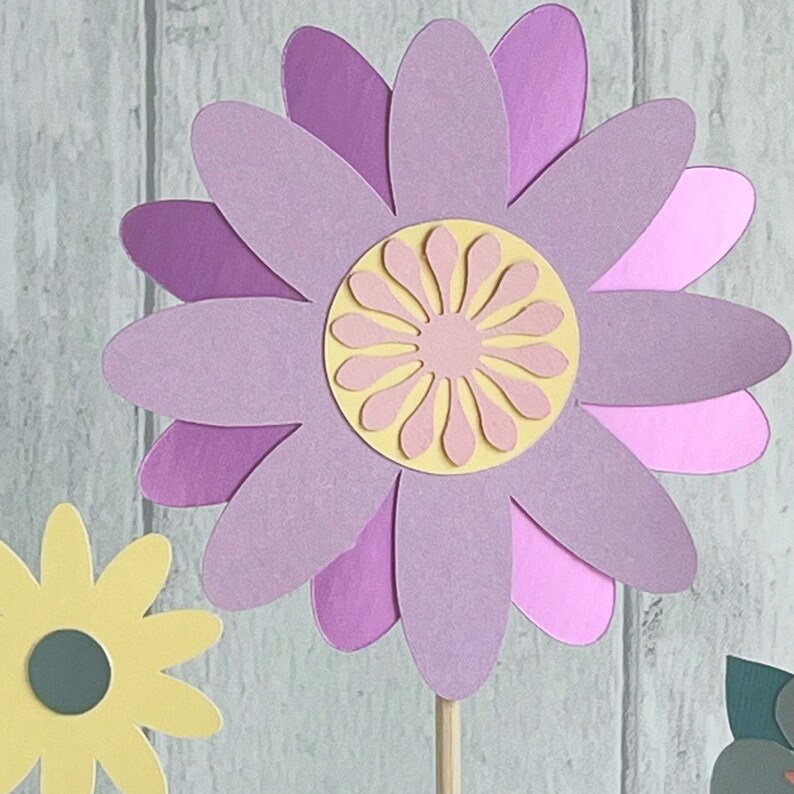 Pu&ograve; includere: Un fiore di carta artigianale con uno schema di colori viola, rosa e giallo. Il fiore ha un design a strati con pi&ugrave; petali.