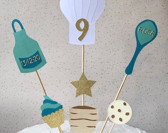 May include: A cake topper set featuring a white chef's hat, a teal apron, a teal spoon with the word "ROSA" on it, a gold star, a teal cupcake, and a stack of pancakes. The chef's hat has the number "9" on it. The apron has the date "13-12-20" on it.