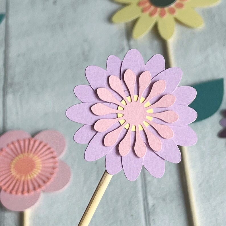 Pu&ograve; includere: Un set di tre topper di fiori di carta in tonalit&agrave; rosa, giallo e viola. I fiori sono sovrapposti e hanno un aspetto delicato e fantasioso. Sarebbero perfetti per aggiungere un tocco primaverile a una torta o ad un altro progetto artigianale.
