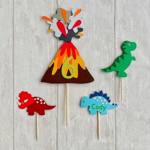 May include: A set of three cake toppers featuring a volcano erupting, a green dinosaur, and a blue dinosaur with the name "Cody" on it. The volcano is brown with orange and yellow flames and a number 6 on it.