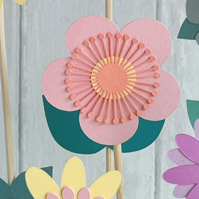 Pu&ograve; includere: Un fiore di carta rosa con un centro giallo e arancione. Il fiore &egrave; attaccato a un bastoncino di legno e ha foglie verdi.