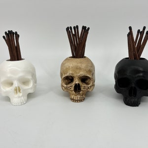 May include: Three skull-shaped matchstick holders, each with a different color: white, beige, and black. Each skull has a pile of brown matchsticks sticking out of the top.