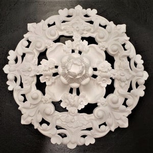 May include: White decorative ceiling medallion with a floral design. The medallion has a central flower with intricate details and is surrounded by a delicate, lacy pattern.