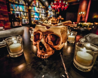 May include: A skull-shaped ashtray with two cigars resting on the rim. The ashtray is surrounded by four lit candles on a dark wooden table.