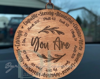 May include: Wooden car charm with the words "You Are" in a script font, surrounded by inspirational words and Bible verses. The charm is round and hangs from a string with wooden and leopard print beads.