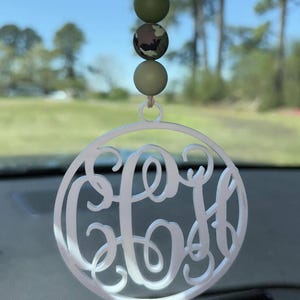 Può includere: Un ornamento circolare bianco con un monogramma composto da tre lettere intrecciate in corsivo. L'ornamento pende da uno spago color cuoio decorato con tre perline rotonde: due perline verde oliva tinta unita e una centrale con motivo mimetico.