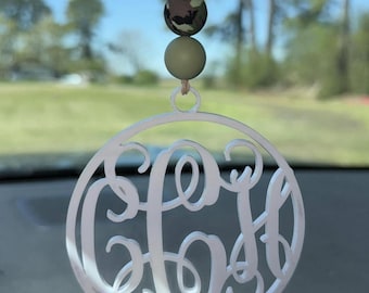 Può includere: Un ornamento circolare bianco con un monogramma composto da tre lettere intrecciate in corsivo. L'ornamento pende da uno spago color cuoio decorato con tre perline rotonde: due perline verde oliva tinta unita e una centrale con motivo mimetico.