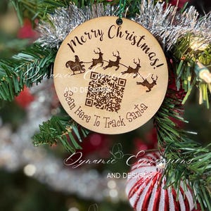 May include: A round wooden Christmas ornament with the words "Merry Christmas!" and "Scan Here To Track Santa" engraved on it. The ornament features a design of Santa's sleigh and reindeer. It hangs on a Christmas tree with other ornaments.