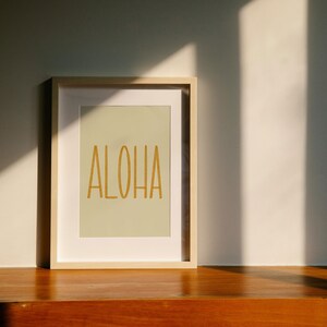 May include: A framed print with a light green background and the word "ALOHA" in gold lettering. The frame is light wood and is sitting on a wooden surface.