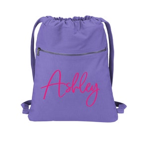 May include: Purple drawstring backpack with a zippered pocket and the name "Ashley" printed in pink.