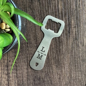 Może przedstawiać: Srebrny otwieracz do butelek z prostokątnym otworem i długim uchwytem. Na uchwycie wygrawerowano litery "L" i "M" oddzielone poziomą linią. Otwieracz leży na drewnianej powierzchni.