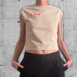 Könnte beinhalten: Beiges Crop-Top mit drei orangefarbenen Punkten auf der linken Brust. Das Oberteil wird mit einer schwarzen Hose kombiniert. Das Outfit ist lässig und modern.