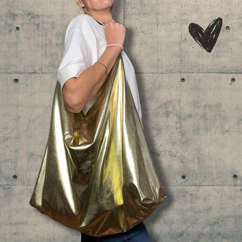 Pode incluir: Uma bolsa tote dourada met&aacute;lica com um design desleixado e sem estrutura. A bolsa &eacute; feita de um material brilhante e reflexivo e tem um topo grande e aberto.