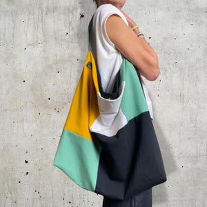 Könnte beinhalten: Eine große, farbenfrohe Tragetasche mit geometrischem Muster. Die Tasche besteht aus vier verschiedenen Farben: Gelb, Weiß, Grün und Schwarz. Die Tasche wird von einer Person getragen, aber das Gesicht der Person ist nicht sichtbar.