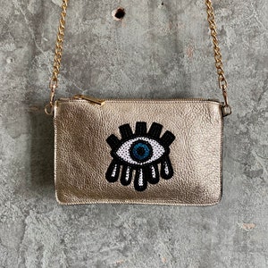 May include: A gold leather clutch bag with a chain strap and a beaded evil eye design.
