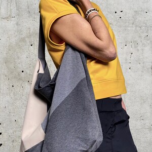 May include: A grey and beige fabric tote bag with a long strap. The bag is worn over the shoulder of a person wearing a yellow shirt and black trousers.