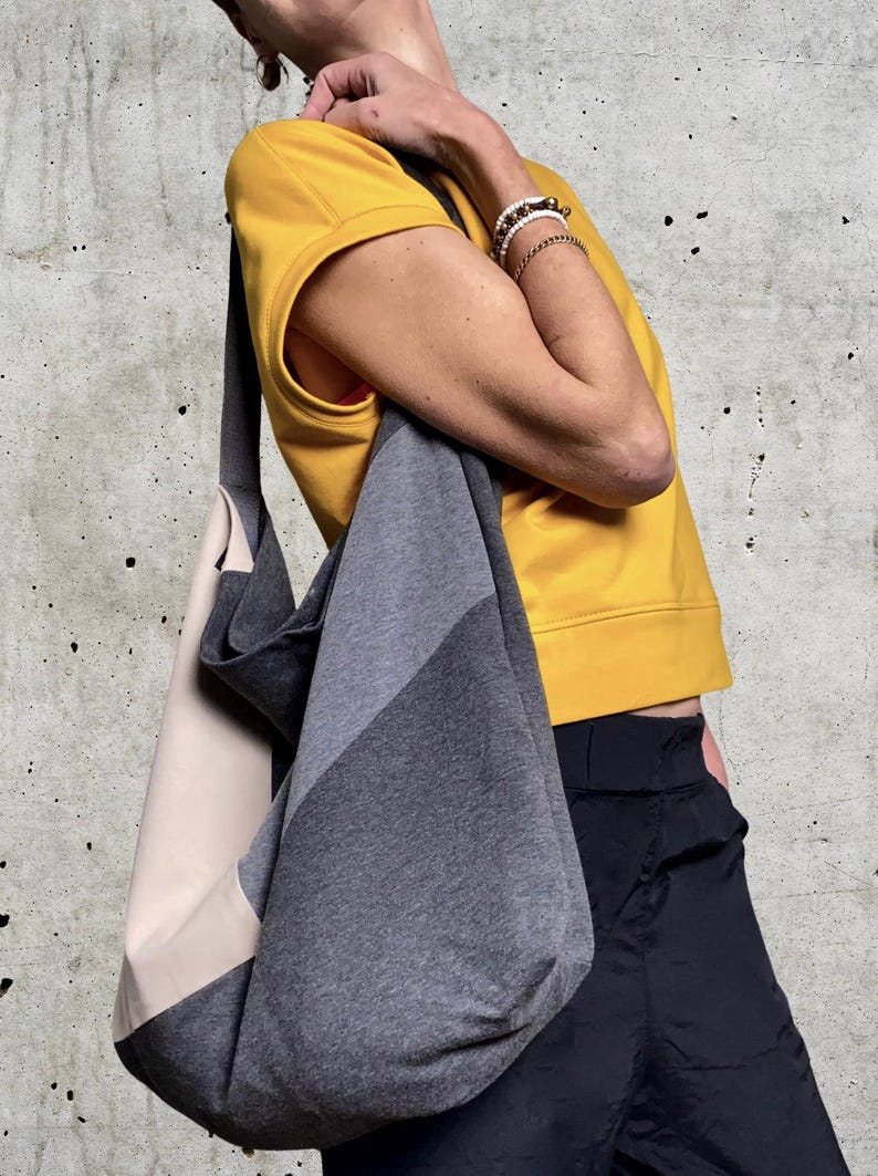 May include: A grey and beige fabric tote bag with a long strap. The bag is worn over the shoulder of a person wearing a yellow shirt and black trousers.