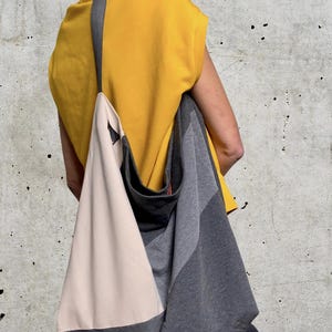 May include: A person wearing a yellow top and black trousers is carrying a large, grey and beige tote bag with a black strap.