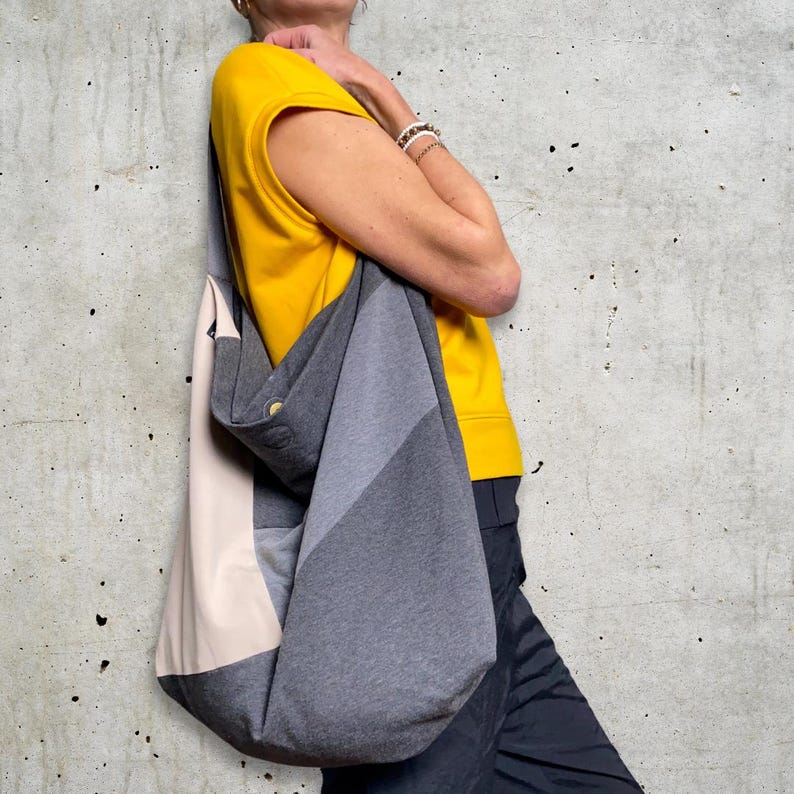 May include: A grey and beige tote bag with a single button closure. The bag is made of a soft, textured fabric and has a wide, open top.