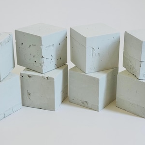 Può includere: Una collezione di nove cubi di cemento grigio chiaro, alcuni impilati, con superfici strutturate e imperfezioni visibili. I cubi sono disposti su una superficie bianca.