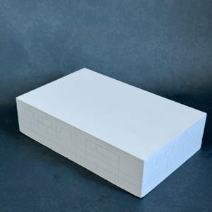 May include: A rectangular, white block with a textured surface. The sides of the block are detailed with a brick pattern, adding a touch of architectural design. The block is set against a dark background, highlighting its clean lines and form.