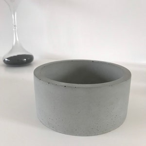 Peut inclure: Un pot de fleur cylindrique en béton gris avec un extérieur lisse. Le pot est de conception simple et moderne, adapté aux petites plantes ou aux objets décoratifs. Un sablier est visible en arrière-plan.