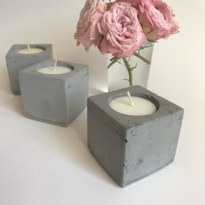 May include: Three gray concrete candle holders with white candles. The candle holders are square shaped and have a rough texture. A bouquet of pink roses is in the background.