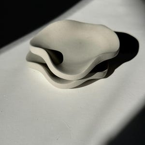 Może przedstawiać: Dwa ułożone na sobie, kremowe ceramiczne talerze o falistym, organicznym kształcie.