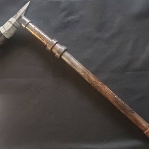 Könnte beinhalten: Eine handgefertigte Spitzhacke im rustikalen Stil mit einem Metallkopf und einem langen, zylindrischen Stiel. Der Kopf hat eine Spitze und eine flache Schlagfläche. Der Stiel besteht aus Metallrohren und -fittings mit einem verwitterten Aussehen.