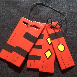 Puede incluir: Un conjunto de cinco etiquetas de madera rojas con detalles en negro y una cuerda negra. Algunas etiquetas presentan diseños de diamantes y círculos amarillos. Las etiquetas están conectadas por una cuerda negra.
