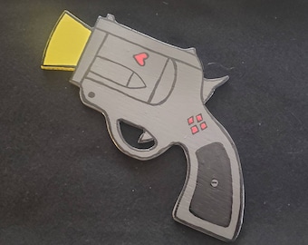 May include: A cartoon-style revolver in grey with black and red details. The gun has a yellow rectangle at the barrel, a red heart, and red diamond shapes. The handle is black.