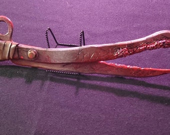 May include: A handcrafted, rustic tool with a loop handle and a scissor-like design. The tool is primarily brown with reddish accents, featuring a wrapped handle and a rivet. The tips have a textured, reddish appearance.
