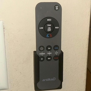 May include: A dark gray Artika remote control for a ceiling fan and light fixture, held in a black wall-mounted plastic holster. The remote features buttons for fan speed, light dimming, timer settings, and color temperature control with clear text labels.