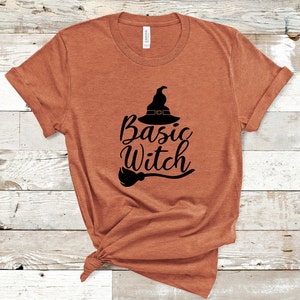 May include: A rust-colored t-shirt with the words "Basic Witch" in black script, accompanied by a witch hat and broom graphic. The shirt is made of a soft material and has rolled-up sleeves.