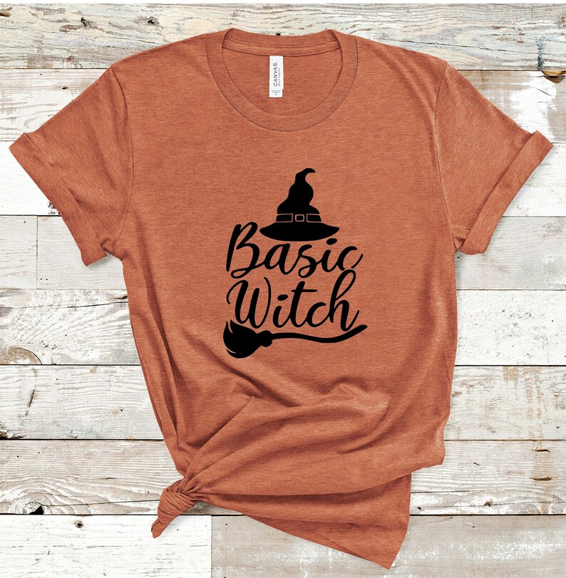 May include: A rust-colored t-shirt with the words "Basic Witch" in black script, accompanied by a witch hat and broom graphic. The shirt is made of a soft material and has rolled-up sleeves.