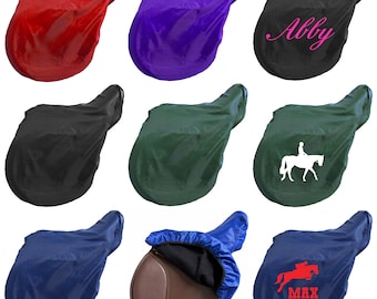 May include: Collection of saddle covers in various colors: red, purple, black, green, navy blue, and a brown saddle with a blue cover. Some covers have text or graphics, including the name "Abby", a rider on a horse, and the name "MAX" with a jumping horse.