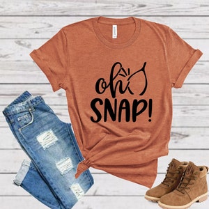 May include: A rust-colored t-shirt with the text "oh snap!" in black lettering. The shirt is paired with blue jeans and brown boots.