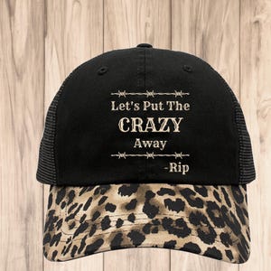 May include: Black and brown trucker hat with a leopard print brim. The hat has a black mesh back and the text "Let's Put The CRAZY Away -Rip" in beige.